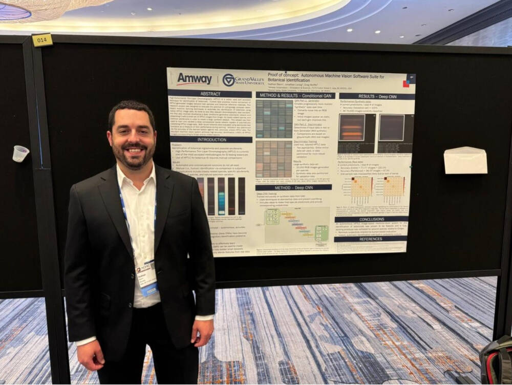 Nathan Stern, in a black suit and white shirt stands in front of a scientific poster presentation. The poster, displayed on a black board labeled "014," features the logos of Amway and Grand Valley State University and is titled "Proof of concept